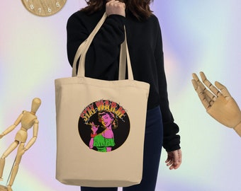 Könnte beinhalten: Eine beige Tragetasche mit einem schwarzen Kreisdesign, das eine Frau mit rosa Haaren und einem grünen Kleid zeigt. Der Text "STAY WARM" ist in gelben und orangefarbenen Buchstaben um den Kreis herum geschrieben.