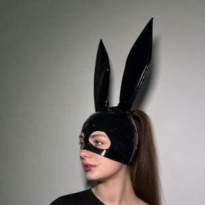 May include: A black glossy latex hood featuring two tall, pointed rabbit ears extending from the top. The headpiece covers the scalp and forehead, with a large cutout opening for the eyes and nose, constructed with visible seams along the surface.