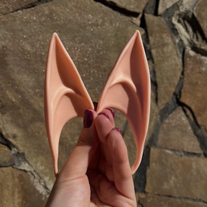 May include: A pair of pale pink elf ears, held up against a stone background. The ears are pointed and curved, with a smooth texture. The ears are designed to be worn, likely for cosplay or costume purposes.