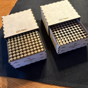 May include: Two wooden boxes with sliding lids, each labeled "223 Rem." One box is open, revealing numerous brass-colored cartridges. The other box is partially open, showing a grid of holes. The boxes are on a dark surface.
