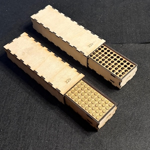 May include: Two wooden boxes with sliding lids, each containing small, brass-colored cylindrical objects. The boxes are made of light-colored wood and have a notched design along the edges. The text "22lr" is visible on the boxes.