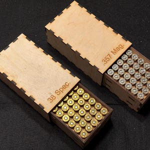 May include: Two rectangular light-colored wooden sliding boxes with finger-jointed edges. One box is engraved with .357 Mag. and contains silver-colored cartridges. The other box is engraved with 38 Spec. and contains brass-colored cartridges.