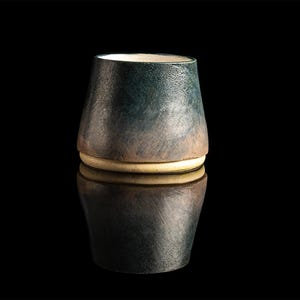 May include: A small, dark green ceramic cup with a light brown rim. The cup is sitting on a black surface and is reflected in the surface.