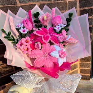May include: A colourful bouquet of handmade flowers in pink, white, and green. The bouquet includes various flower types, wrapped in pink tissue paper and tied with a sparkly white ribbon. The flowers are made of soft, textured materials.