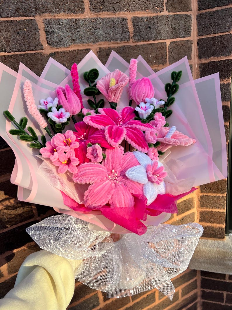 May include: A colourful bouquet of handmade flowers in pink, white, and green. The bouquet includes various flower types, wrapped in pink tissue paper and tied with a sparkly white ribbon. The flowers are made of soft, textured materials.