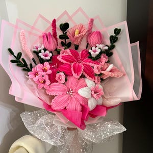 Puede incluir: Un ramo de flores rosas hechas a mano, que incluye lirios, tulipanes y otras flores, envuelto en papel de seda rosa y atado con una cinta blanca brillante. Las flores están hechas de un material suave y texturizado.