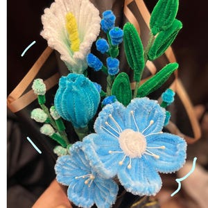 May include: A handmade bouquet of pipe cleaner flowers in shades of blue, white, and green. The bouquet includes a large white calla lily with a yellow center, blue flowers, and green leaves, wrapped in black paper.