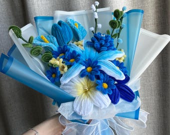 May include: A handcrafted bouquet of blue and white flowers, presented in blue and white wrapping and tied with a white ribbon. The bouquet includes various shades of blue flowers with yellow centres and green accents.