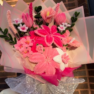 May include: A handcrafted bouquet featuring pink and white flowers, such as lilies and tulips, with green stems. The bouquet is wrapped in pink and translucent paper and tied with a silver, glittery mesh. The flowers are made of fabric.