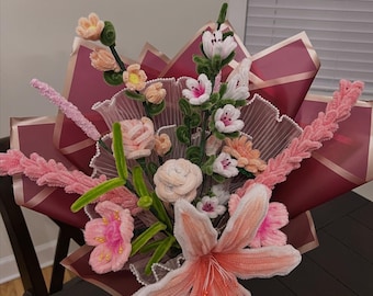 May include: A bouquet of handmade flowers in shades of pink, peach, and white, wrapped in burgundy and pink paper. The arrangement includes lilies, roses, and other floral designs, with green stems and accents. The bouquet is displayed on a dark brown table.