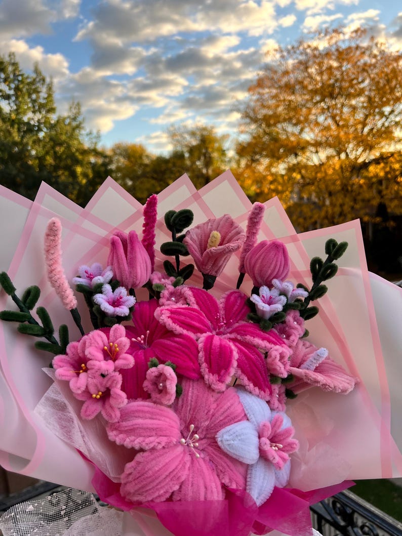May include: A bouquet of handmade pink flowers, including lilies, tulips, and smaller blossoms, wrapped in pink and white paper. The flowers are made of a soft, textured material. The background features a sky with clouds and trees.
