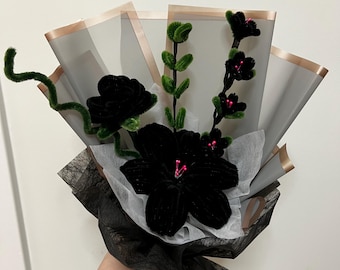 May include: A handcrafted bouquet featuring black velvet flowers with green stems and accents. The arrangement is wrapped in layers of grey and black tissue paper, with gold trim. A distinctive and artistic floral display.