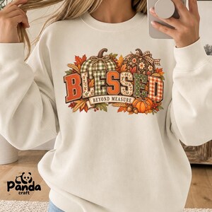 May include: A cream-colored crewneck sweatshirt featuring a graphic print of pumpkins, autumn leaves, and wheat. The text BLESSED BEYOND MEASURE is displayed in large, patterned block letters across the center, accented by small acorns.
