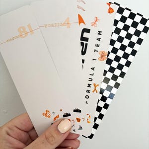 May include: A hand holding a stack of Formula 1 themed bookmarks. The bookmarks feature racing car illustrations, the text "Formula 1 Team", and a checkered flag design. The bookmarks have the names "Piastri" and "Norris" with the numbers 81 and 4.