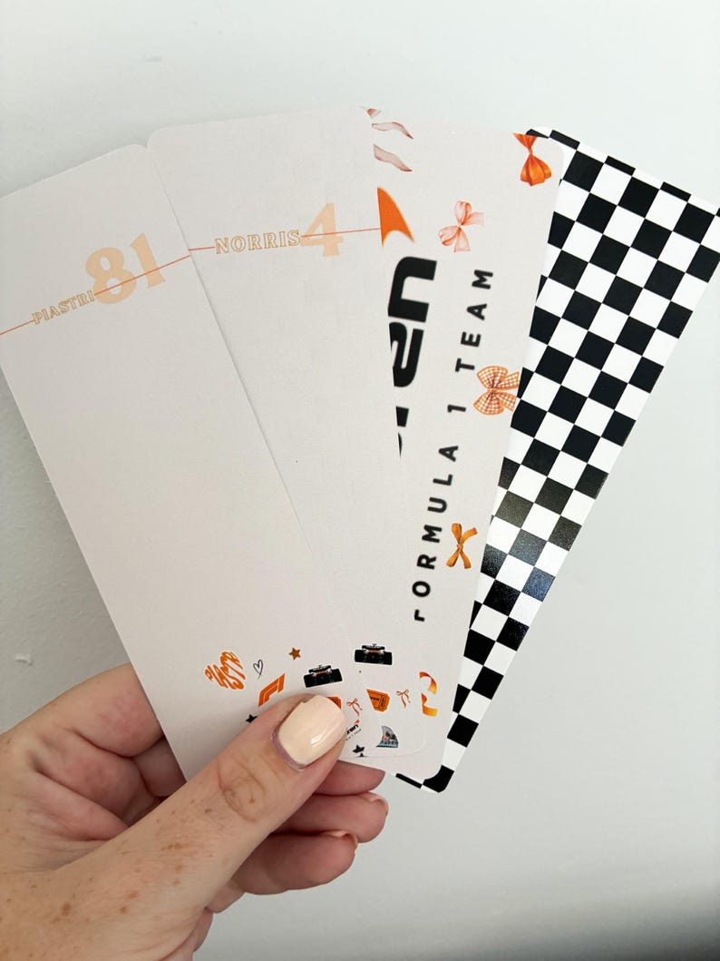 May include: A hand holding a stack of Formula 1 themed bookmarks. The bookmarks feature racing car illustrations, the text "Formula 1 Team", and a checkered flag design. The bookmarks have the names "Piastri" and "Norris" with the numbers 81 and 4.