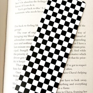 May include: A rectangular bookmark with a black and white checkered pattern. The bookmark is positioned over an open book, with text from the book visible in the background. The design is a classic, simple pattern.