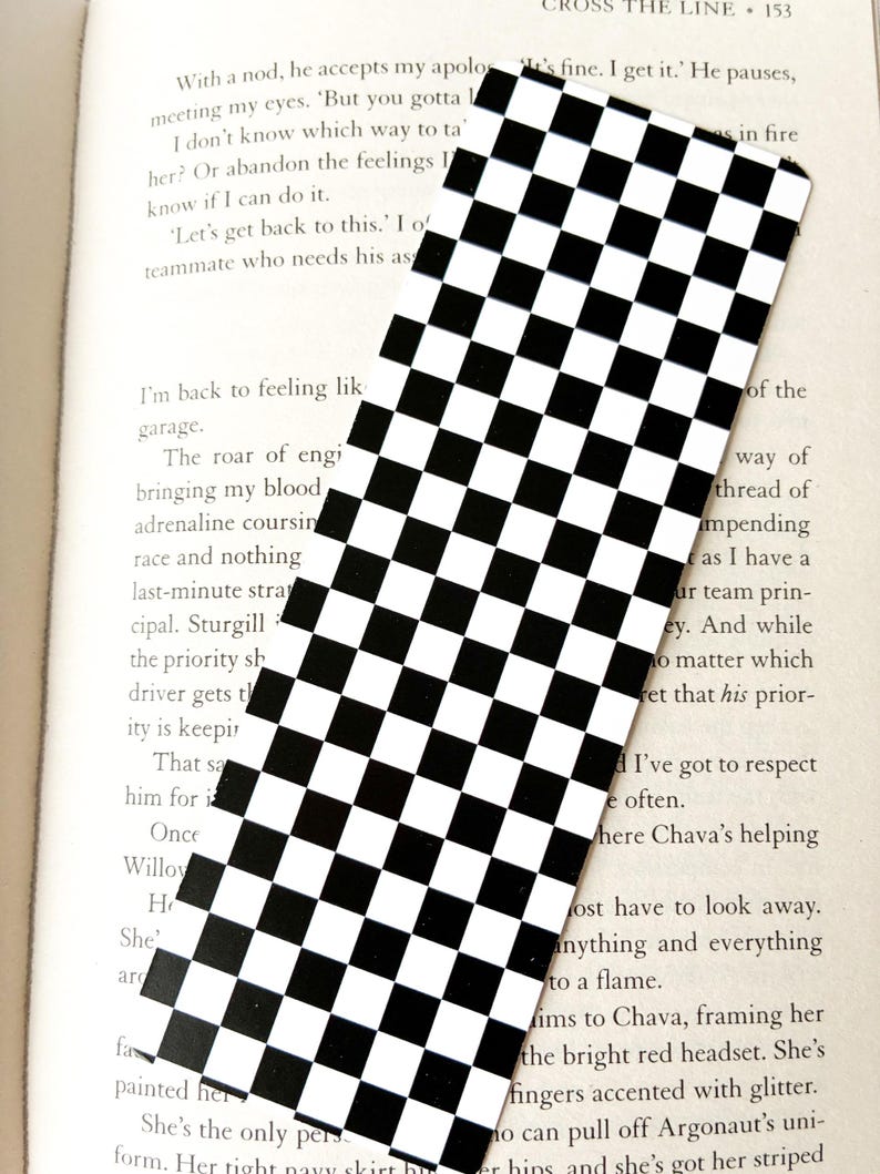 May include: A rectangular bookmark with a black and white checkered pattern. The bookmark is positioned over an open book, with text from the book visible in the background. The design is a classic, simple pattern.