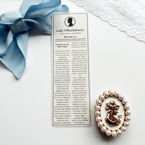 May include: A light blue ribbon bow, a bookmark with text, and a gold-colored cameo brooch with faux pearls. The bookmark features the text "Lady Whistledown's Society Papers". The cameo brooch has a woman's silhouette.