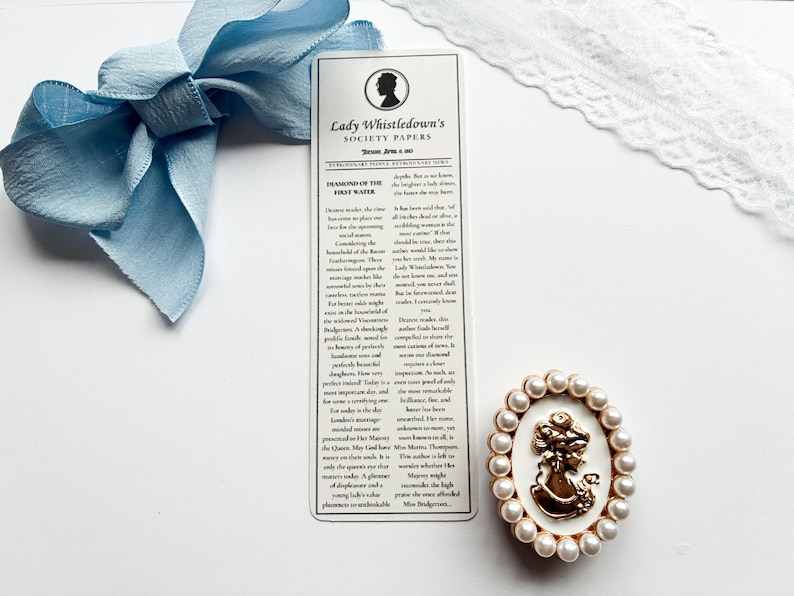 May include: A light blue ribbon bow, a bookmark with text, and a gold-colored cameo brooch with faux pearls. The bookmark features the text "Lady Whistledown's Society Papers". The cameo brooch has a woman's silhouette.