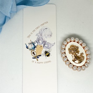 May include: A white bookmark with a whimsical illustration and the text "THIS IS THE FIRST CHAPTER OF A HAPPY ENDING." A gold and white cameo brooch with pearl accents is also shown. A light blue ribbon is in the background.