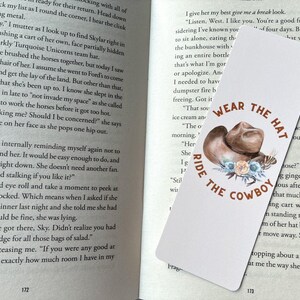 May include: A white bookmark featuring the text "WEAR THE HAT RIDE THE COWBOY" in brown. The design includes a brown cowboy hat, floral accents, and feathers. The bookmark is on an open book.