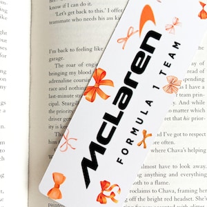 May include: A white McLaren Formula 1 Team bookmark with the team's logo in black and orange. The bookmark is decorated with watercolor-style orange bows. The bookmark is placed on top of an open book.