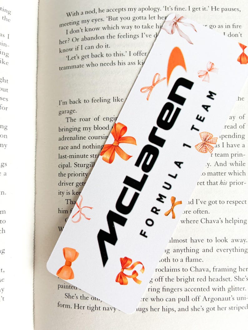 May include: A white McLaren Formula 1 Team bookmark with the team's logo in black and orange. The bookmark is decorated with watercolor-style orange bows. The bookmark is placed on top of an open book.