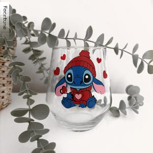 May include: A stemless wine glass featuring a cartoon character in a red hat and sweater, with red hearts scattered around. The character is blue with large eyes and pink ears. The glass is clear, and the design is printed on the front.