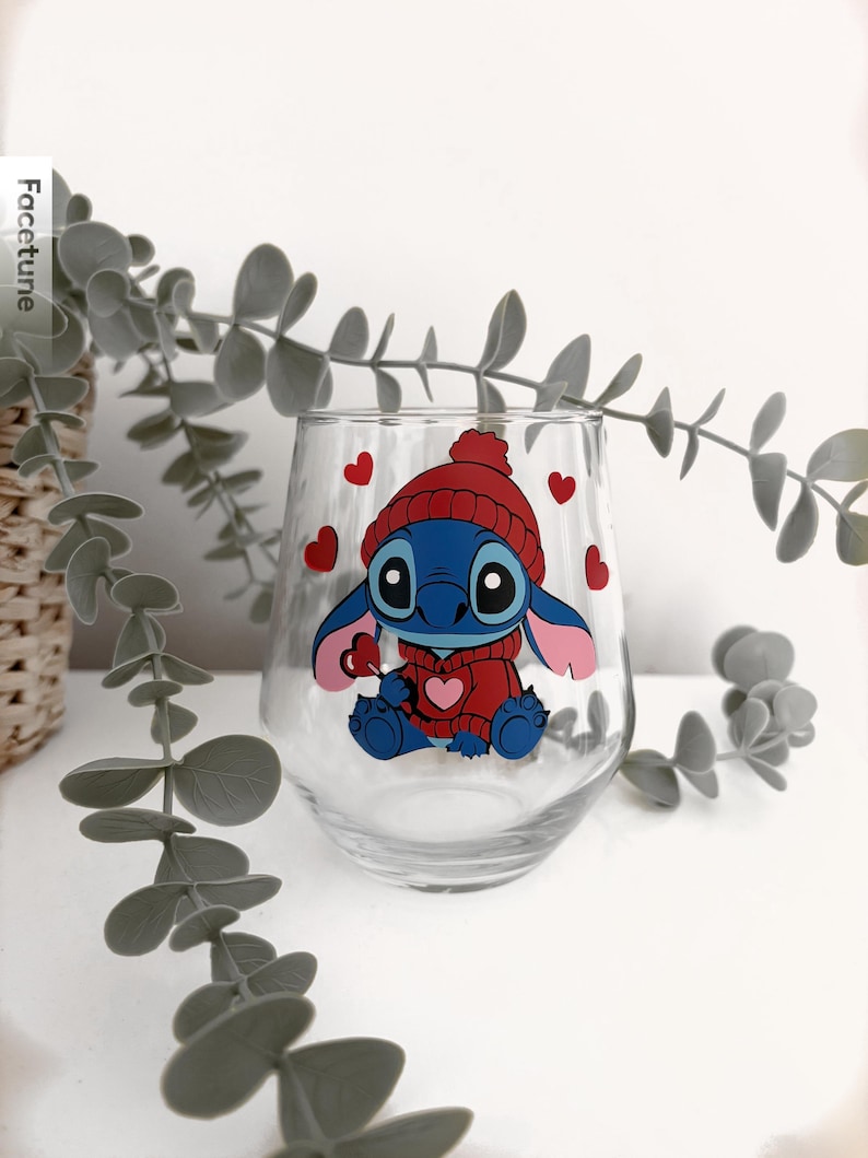 May include: A stemless wine glass featuring a cartoon character in a red hat and sweater, with red hearts scattered around. The character is blue with large eyes and pink ears. The glass is clear, and the design is printed on the front.