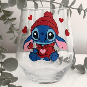 May include: A clear stemless wine glass featuring a blue cartoon character wearing a red hat and sweater, holding a heart-shaped lollipop. Red hearts are scattered around the character. The glass is decorated with a colorful, whimsical design.