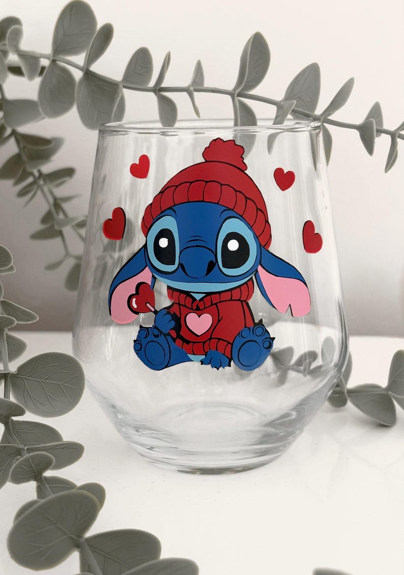 May include: A clear stemless wine glass featuring a blue cartoon character wearing a red hat and sweater, holding a heart-shaped lollipop. Red hearts are scattered around the character. The glass is decorated with a colorful, whimsical design.