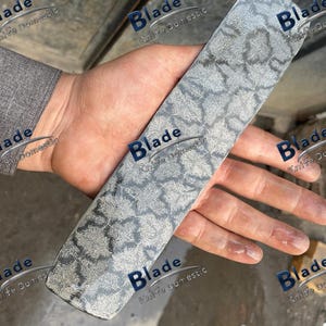 May include: A rectangular blade with a floral pattern. The blade is held in a hand, and the text "Blade Knife Domestic" is visible. The blade's surface has a textured, grey and white pattern. The blade is likely made of metal.