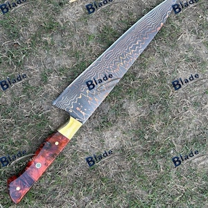May include: A large chef's knife with a patterned blade and a wooden handle. The blade has a wavy design in shades of brown and gold. The handle is a mix of red and brown wood with brass accents.