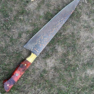 May include: A large chef's knife with a patterned blade and a wooden handle. The blade has a wavy, textured design with copper-colored accents. The handle is a combination of red, brown, and gold wood, with decorative rivets.