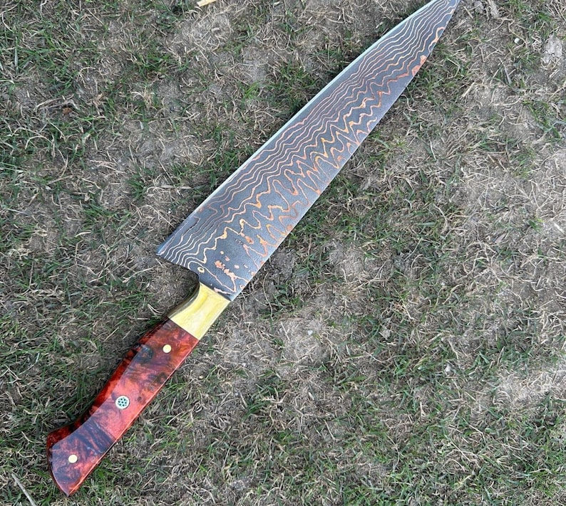 May include: A large chef's knife with a patterned blade and a wooden handle. The blade has a wavy, textured design with copper-colored accents. The handle is a combination of red, brown, and gold wood, with decorative rivets.