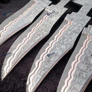 Könnte beinhalten: Nahaufnahme von fünf Stahlmesserklingen mit einem Muster. Die Klingen haben ein wellenförmiges, geschichtetes Muster in Grau- und Weißtönen mit einem rosafarbenen Akzent. Auf jeder Klinge ist das Wort "Blade" aufgedruckt.