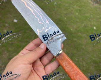 May include: A handmade chef's knife with a patterned steel blade and a reddish-brown handle. The blade has a unique design with copper-colored swirls. The handle is a solid, textured material. The knife is designed for kitchen use.