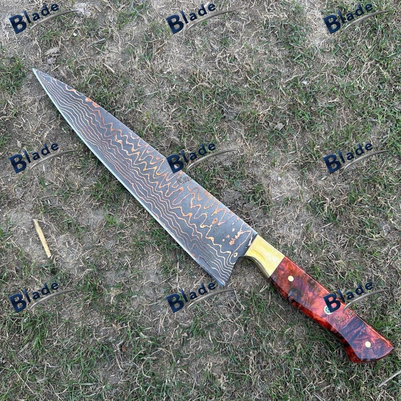 May include: A large, Damascus steel chef's knife with a patterned blade and a red and brown wooden handle. The handle has a brass bolster. The knife is lying on a bed of grass.