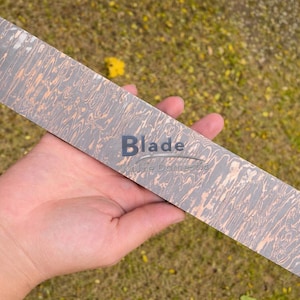 Puede incluir: Una hoja de cuchillo rectangular con un diseño estampado en tonos grises y cobrizos se sostiene en una mano. La palabra "Blade" está impresa en la hoja. La hoja tiene una apariencia texturizada única.