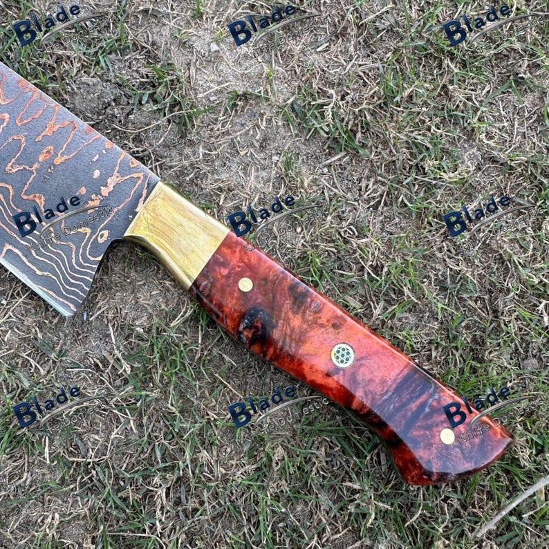 May include: A Damascus steel chef's knife with a red and brown burl wood handle. The blade has a unique pattern, and the handle is accented with brass and small rivets. The knife is designed for kitchen use.