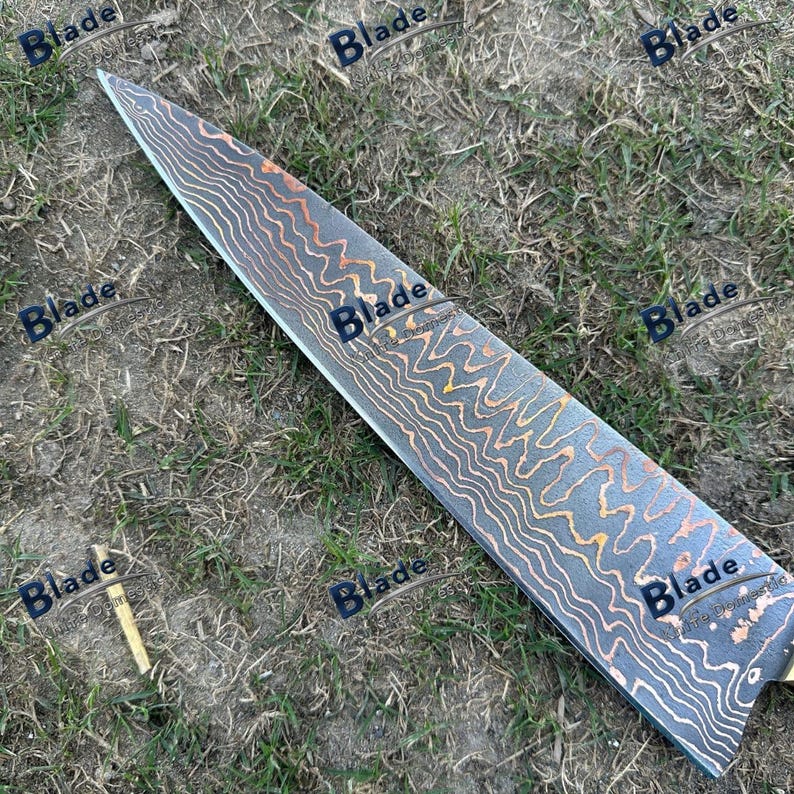 May include: A long, Damascus steel blade with a distinctive pattern of swirling dark and copper tones. The blade is set against a backdrop of green grass, showcasing its intricate design and sharp edge. The blade has a brass handle.