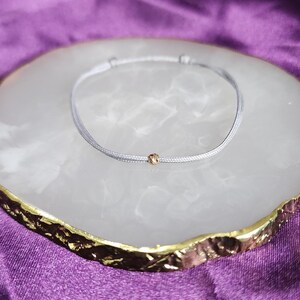 May include: A delicate grey bracelet with a small gold bead at the centre. The bracelet is displayed on a white and gold decorative tray, set against a purple background. The bracelet is simple and elegant.