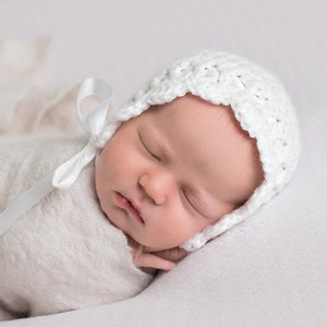 May include: A white crocheted baby bonnet with a white satin ribbon tied under the chin. The bonnet has a scalloped edge and is worn by a sleeping baby.