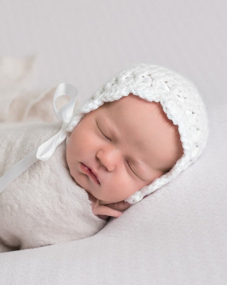 May include: A white crocheted baby bonnet with a white satin ribbon tied under the chin. The bonnet has a scalloped edge and is worn by a sleeping baby.