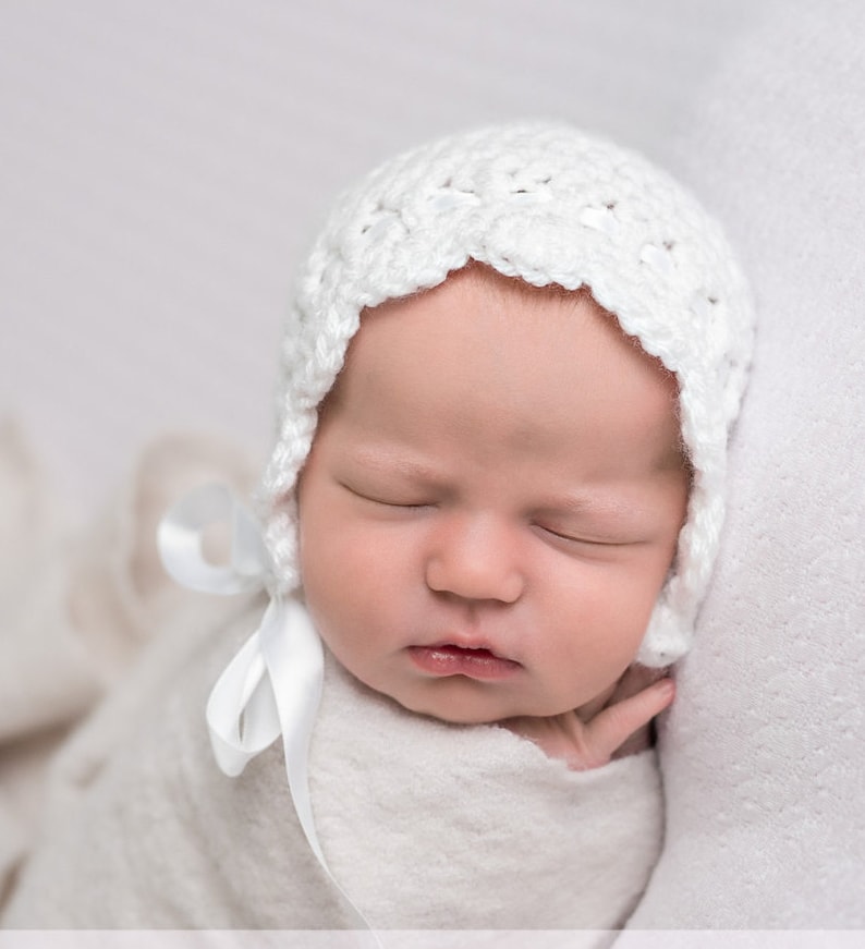 May include: A sleeping newborn baby wearing a white crocheted bonnet with a white satin ribbon tied under the chin.
