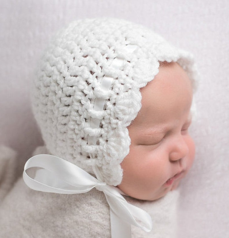 May include: A white crocheted baby bonnet with a white satin ribbon tied at the back. The bonnet has a scalloped edge and a simple design.