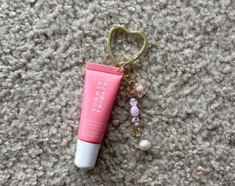 Puede incluir: Un tubo de bálsamo labial rosa con el texto "SUMMER FRIDAYS" y "LIP BUTTER BALM" en la parte delantera. El tubo está unido a un llavero de corazón dorado con abalorios de caramelo rosa y blanco y una perla.
