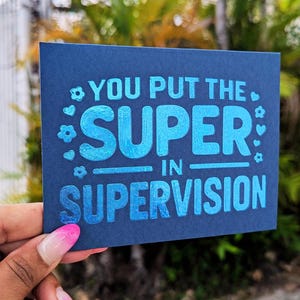May include: A navy blue card with the text "YOU PUT THE SUPER IN SUPERVISION" in bright blue, with decorative floral and heart accents. The card is held in a hand with a pink and white manicure.