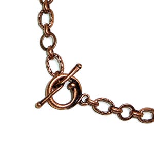 May include: Close-up of a copper-colored necklace with an oval link chain and a toggle clasp. The chain links have a textured, hammered appearance. The toggle clasp is a circular design with a bar closure.