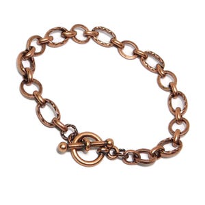 May include: A copper-colored bracelet with an oval and round link chain design. The bracelet features a toggle clasp closure. The links have a textured pattern. The jewelry is displayed on a white background.